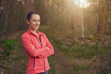 Happy confident young woman with a friendly smile standing with folded arms in woodland backlit by the glow of the setting sun enjoying the spring weather