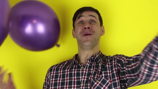 A funny, cute guy is actively playing with a purple balloon. A portrait of a young guy, which actively expresses joy and plays with a purple balloon. A portrait on a yellow background.