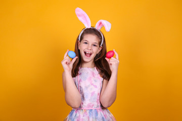 Obraz na płótnie Canvas Studio portrait of smiling young girl wearing traditional bunny ears headband for easter. Brunette female with pigtails holding traditional colored eggs over yellow background. Close up, copy space.