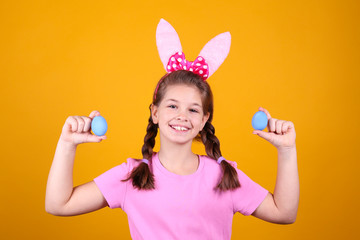 Studio portrait of smiling young girl wearing traditional bunny ears headband for easter. Brunette female with pigtails holding traditional colored eggs over yellow background. Close up, copy space.