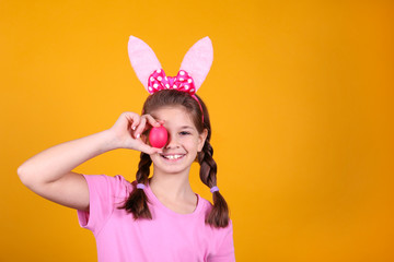 Studio portrait of young girl wearing traditional bunny ears headband for easter, covering her eyes with an egg and smiling. Brunette female with pigtails over yellow background. Close up, copy space.