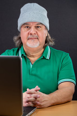 Man poses working on laptop at his desk. He is in his 50s. He is wearing a t-shirt and a hood. He...