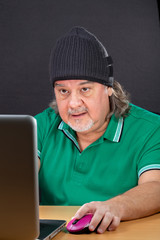 Man working on laptop at his desk. He is in his 50s. He is wearing a t-shirt and a hood. He has long hair. This is a studio image. He is deeply concentrated on his work.