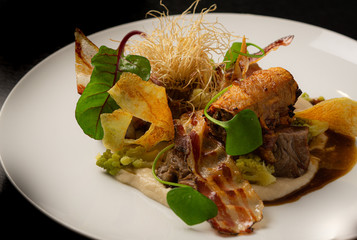 Roasted pork belly with potato puree, bacon, sauce, vegetables, and green salad leaves served on white plate on black background. Restaurant dish. 