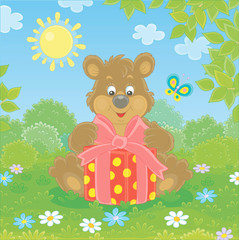 Obraz na płótnie Canvas Friendly smiling little brown bear holding a beautiful box with a holiday gift on green grass of a pretty forest glade on a sunny summer day, vector cartoon illustration