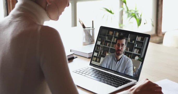 Over shoulder view of woman student worker makes conference video call on laptop talks with online teacher, boss or partner in web cam chat on computer screen. Distance work, remote education concept.
