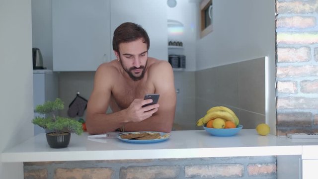 Serious, shirtless, fit man looking at his mobile phone while leaning on the kitchen counter with protein crepes on. Pan shoot from right to left