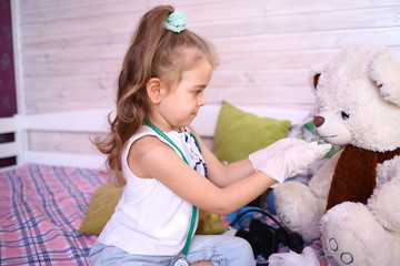 Quarantined fun. Caucasian cute little girl in a medical gloves at home playing doctor with a bear, give pills, treat. Quarantine protection against coronavirus 2019-ncov. Home insulation