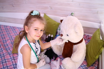 Quarantined fun. Caucasian cute little girl in a medical gloves at home playing doctor with a bear, give pills, treat. Quarantine protection against coronavirus 2019-ncov. Home insulation