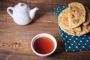 Traditional chebureks on a plate, a bowl filled with hot tea, and a white teapot on a wooden table, top view