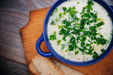 A blue cup with cheese, vegetable soup stands on a cutting, wooden board. Next to the soup with herbs, two slices of bread