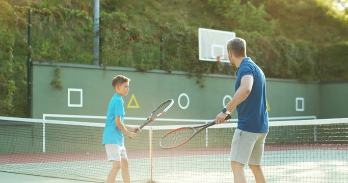 Rear on Caucasian young man teaching his son how to hit ball with racket and giving five on sunny summer day. Back view on male instructor showing to boy how to play tennis on sport court.