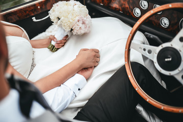 Young married couple sitting in car holding hands and bouquet of flowers, ceremony wedding day, wedding rings
