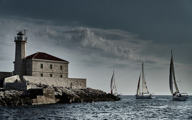 Sailboats compete in a sailing regatta at sunset, sailing race, reflection of sails on water, multi-colored spinnaker, boat number aft boats, big white clouds, the island with a lighthouse