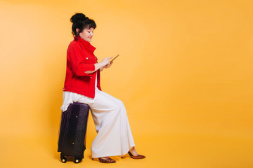 A woman in a red jacket sits on a purple suitcase on a yellow background, holds a smartphone in her hands. Studio photo