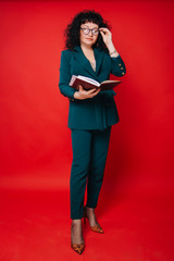 A woman with black curly hair on a red background holds a notebook and pen in her hands. A woman of fifty years in a green suit and glasses.