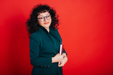 A woman with black curly hair on a red background holds a notebook and pen in her hands. A woman of fifty years in a green suit and glasses.