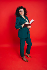 A woman with black curly hair on a red background holds a notebook and pen in her hands. A woman of fifty years in a green suit and glasses.