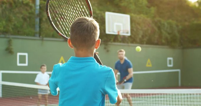 Rear of Caucasian teen boy playing big tennis with joyful handsome father and small sister outdoors in summer. Back view on teenage play with dad and girl at sport court. Family weekend.