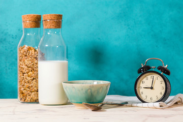 Glass bottles with muesli and milk. On the table is an empty bowl, a spoon and a black alarm clock with a towel