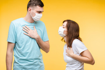 Stock studio of young couple in aseptic masks holding their hands on breast showing that it is hard to breath. COVID-19 symptoms. Pneumonia concept.