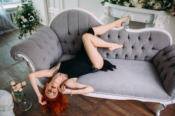 Sexy red-haired young woman with a tattoo is lying on the sofa. Long bare legs - 335381492