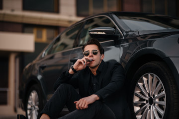 A stylish businessman smokes cigars near a luxury car. Fashion and business