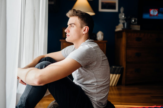 Young man staying at home,looking trough the window.Depressed man avoiding social contact.Relaxing at home.Quarantine self isolation concept.Worried person indoors activity.Anxiety.Agoraphobia