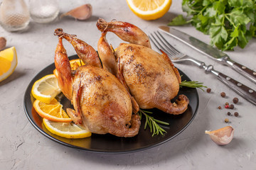 Baked quails with lemon and orange served on a dark plate on a gray background