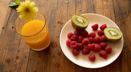 A glass of orange juice and a plate of kiwi with raspberries on a wooden wet background.
