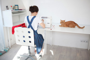 Obraz premium Happy attractive young woman seamstress sitting and sews on sewing machine in studio. Work from home. Remote work during a quarantine. Home concept. Self isolation.