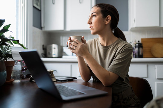 Thoughtful female soldier drinking coffee at laptop in morning kitchen