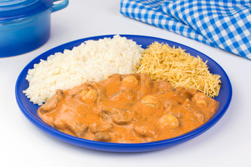 Brazilian tenderloin stroganoff with rice and potato sticks in a blue plate in white background