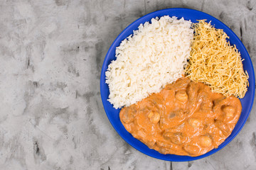 Brazilian tenderloin stroganoff with rice and potato sticks in a blue plate isolated in burnt cement background seen from above