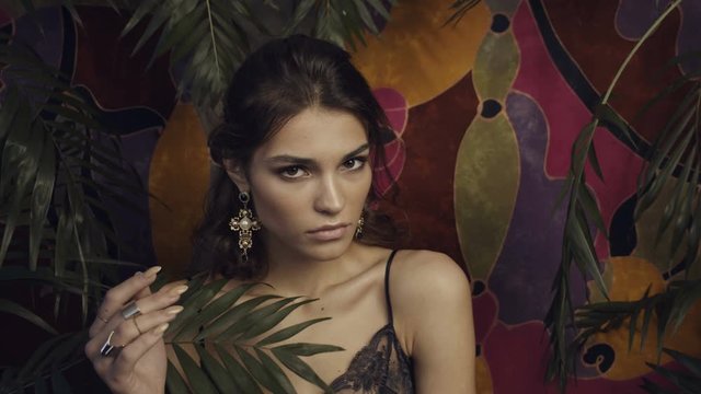 Tanned beautiful girl model with large gold earrings on a motley background among tropical leaves.