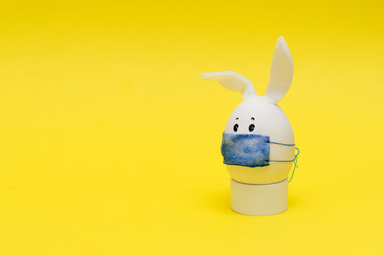 Easter egg with eyes,rabbit ears and a protective mask on the yellow background. Coronavirus epidemic concept. Concept celebrating Easter in self-isolation and quarantine