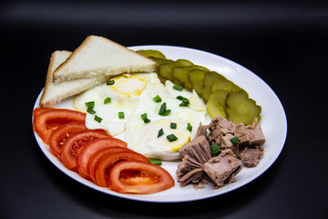 Delicious fresh tuna with eggs, bread, tomatoes, cucmbers with green onion at the morning. Healthy and diet breakfast with tuna, eggs and vegetables on the white plate against dark background