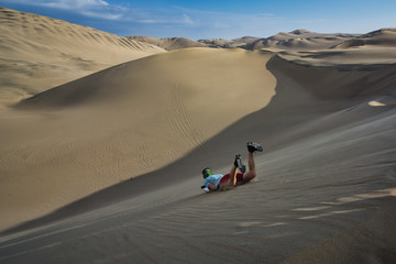 Fototapeta na wymiar Man sandboarding down a large sand dune in the oasis of Huacachina, Peru. Typical, adventurous day activity in the beautiful pacific coastal desert with views of the surreal sandy landscape