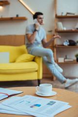 Selective focus of coffee table with papers, cup of coffee and notebook with freelancer talking on smartphone on sofa