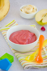 Baby food. Homemade apple puree or sauce with banana and raspberries in a bowl with a spoon on a light background. Healthy food. Vertical orientation.