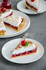 Tart with cherry filling and Italian meringue with a cocktail cherry, chocolate and mint on top on a white plate on a dark concrete background. Cherry pie. Vertical orientation