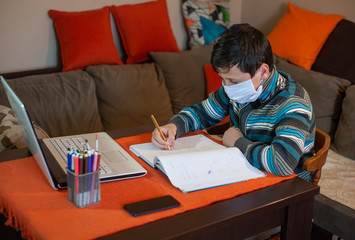 School boy with protective mask in home schooling using laptop during self isolation at her bedroom, online education, doing homework