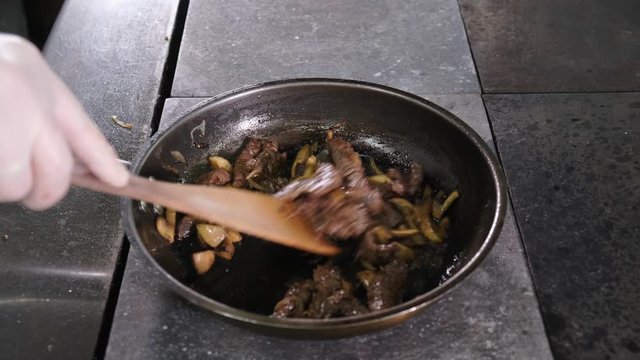 Cook roasts deer meat with onions and mushrooms on hot pan in restaurant kitchen. Chef is stirring and tossing ingredients in pan.