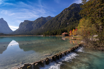 Italian Dolomites, Lago di Dobbiaco - Toblacher See -
calm blue-green water of lake in afternoon sun with shores covered with trees. On the right, water falls over a wooden weir.