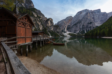 Fototapeta na wymiar Italian Dolomites, Lago di Braies calm, blue-green lake surface with tall shores covered with trees. In the left wooden railing leading to the house which stands on wooden stilts in the water. 