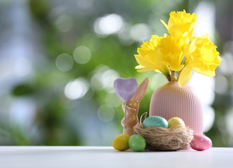 Painted Easter eggs, ceramic bunny and flowers on white table against blurred background. Space for text