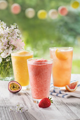 three glasses with fresh juice from passion fruit, strawberries, red orange on a table in the garden, with ice