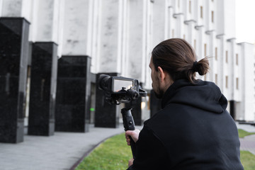 Young Professional videographer holding professional camera on 3-axis gimbal stabilizer. Pro equipment helps to make high quality video without shaking. Cameraman wearing black hoodie making a videos.