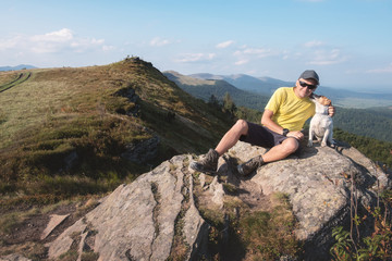 Alone tourist sitting on the edge of cliff with white dog against the backdrop of an incredible mountain landscape. Sunny day and blue sky