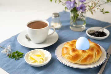 Plaited bread nest with an Easter egg, coffee, butter, jam and flowers on a blue napkin and a white table, festive holidays breakfeast for a single, copy space